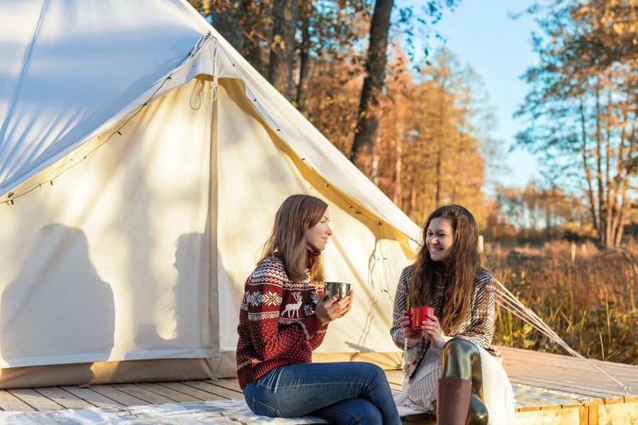 Two happy friends drinking coffee while relaxing near canvas bell tent